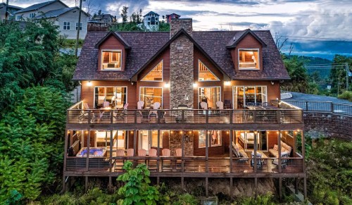A large wooden cabin with a stone facade, featuring multiple balconies and large windows, surrounded by greenery.