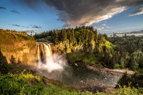A stunning waterfall cascades into a river, surrounded by lush greenery and dramatic clouds in the sky.