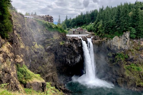 A waterfall cascades down rocky cliffs, surrounded by lush greenery and a cloudy sky, with a building in the background.