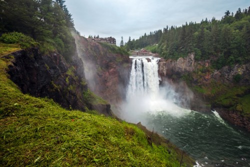 A majestic waterfall cascades into a river, surrounded by lush greenery and mist, with a building visible in the background.