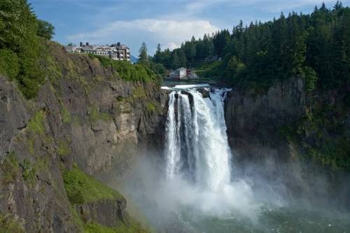 A majestic waterfall cascades down rocky cliffs, surrounded by lush greenery and a distant hotel.