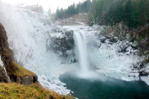 A frozen waterfall cascades into a icy pool, surrounded by rocky cliffs and evergreen trees.