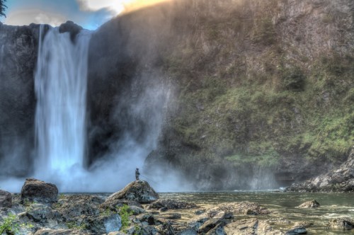 A person stands on a rock near a majestic waterfall, surrounded by mist and lush greenery.