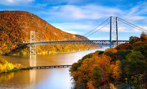 A scenic view of a bridge over a river, surrounded by vibrant autumn foliage and rolling hills under a blue sky.