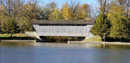 A wooden covered bridge spans a calm lake, surrounded by trees with autumn foliage.