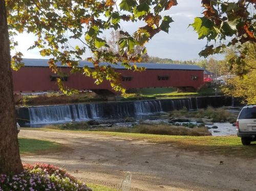 A covered bridge over a waterfall, surrounded by trees and a gravel path, with a vehicle parked nearby.