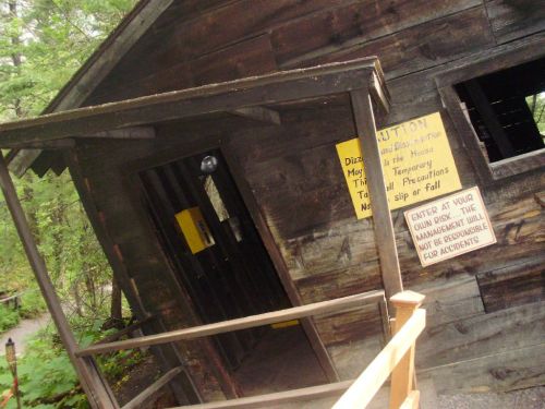 A tilted wooden cabin with warning signs about safety and precautions, surrounded by trees.