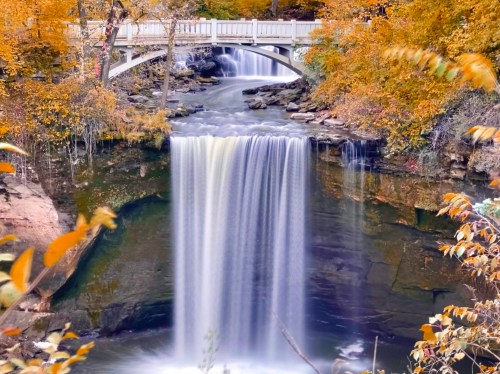 A serene waterfall cascades into a rocky pool, framed by autumn foliage and a white bridge above.