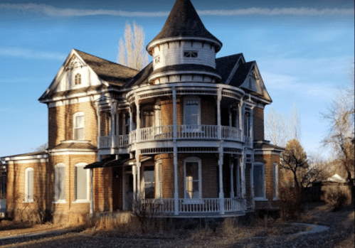 A large, abandoned Victorian-style house with a turret and wraparound porch, surrounded by overgrown vegetation.