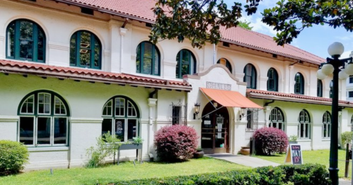 Historic building with arched windows, a red tile roof, and a welcoming entrance surrounded by greenery.