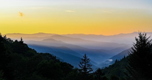 A serene mountain landscape at sunset, featuring layered hills and a gradient sky from orange to blue.