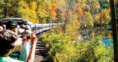 A train travels along a scenic route with colorful autumn foliage, while onlookers take photos by the riverbank.