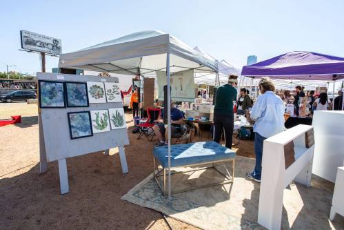 A vibrant outdoor market scene with art displays, tents, and people browsing various booths under a clear blue sky.