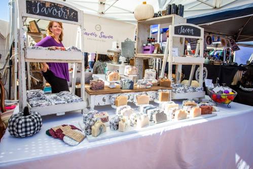 A market stall displays various handmade soaps, with a vendor smiling in the background. Decor includes a small pumpkin.