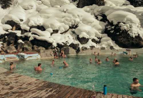 People enjoying a warm pool surrounded by snow-covered rocks and a winter landscape.