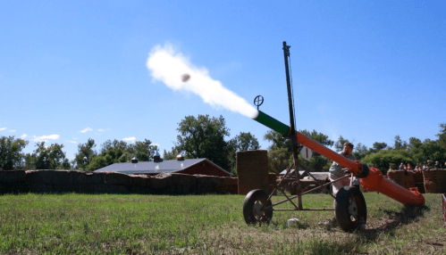A cannon fires a projectile into the sky, with smoke billowing from the barrel against a clear blue backdrop.