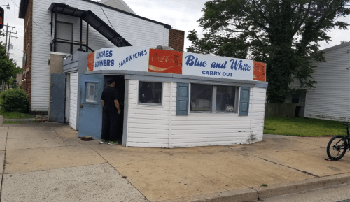 A small carry-out restaurant with a blue and white sign, featuring a window and a door, located on a street corner.