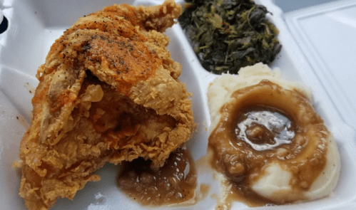 A takeout container with fried chicken, collard greens, mashed potatoes, and gravy.