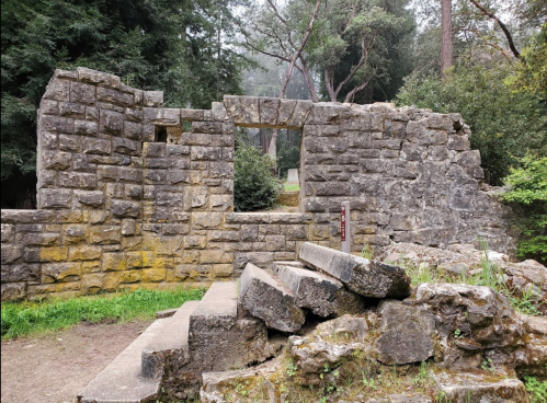 Ruins of a stone structure with an archway, surrounded by trees and greenery. Steps lead up to the entrance.