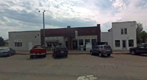 A small town street scene featuring a row of buildings and parked cars under a cloudy sky.