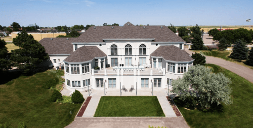 A large, elegant white mansion with a landscaped yard and multiple balconies, set against a clear blue sky.