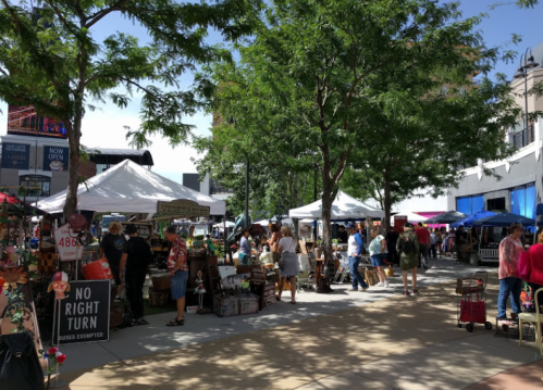 A bustling outdoor market with vendors under white tents, surrounded by trees and shoppers enjoying the day.