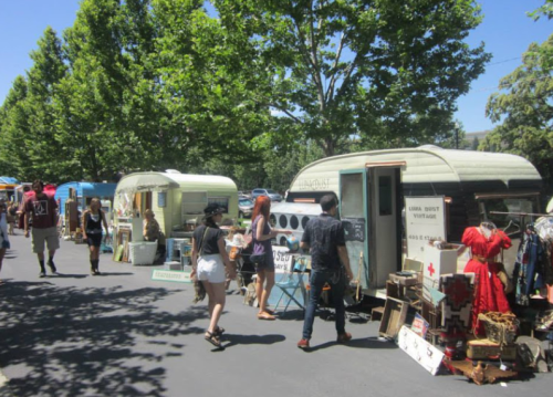 A vibrant market scene with vintage trailers, people browsing, and various items for sale under sunny trees.