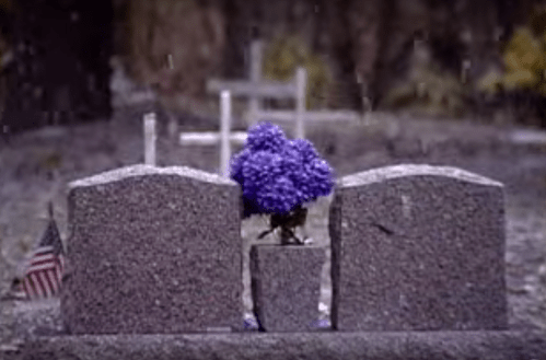 Two gray gravestones with a purple flower arrangement in the center, set against a rainy backdrop and an American flag nearby.
