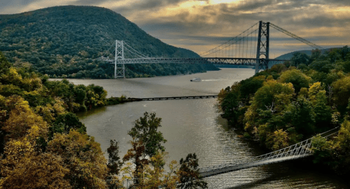 A scenic view of a river with bridges, surrounded by lush green hills and autumn foliage under a cloudy sky.