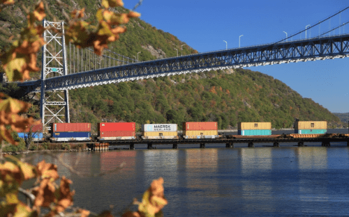 A train with colorful shipping containers travels along a river under a tall bridge, surrounded by autumn foliage.