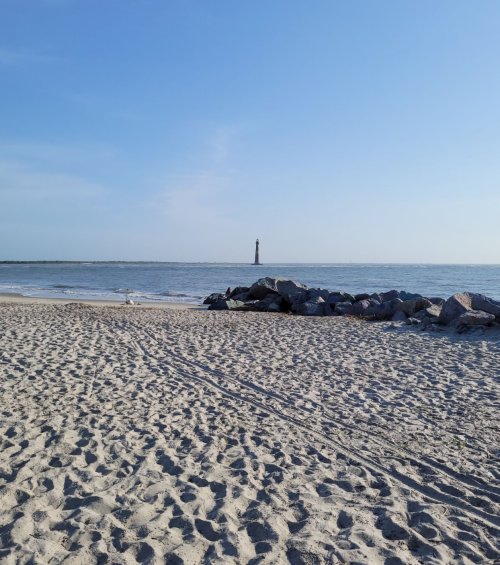 A sandy beach with rocks in the foreground and a lighthouse visible in the distance under a clear blue sky.