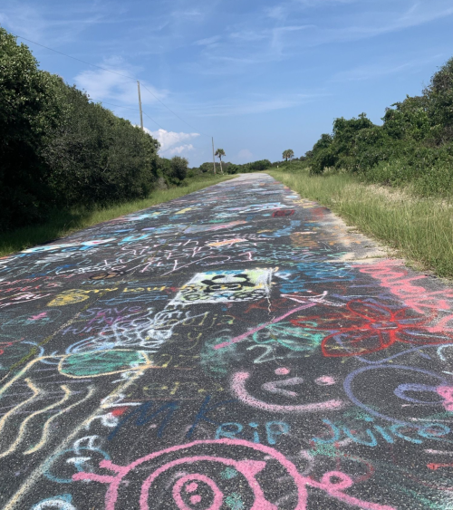 A colorful, graffiti-covered road stretches into the distance, surrounded by greenery and a clear blue sky.