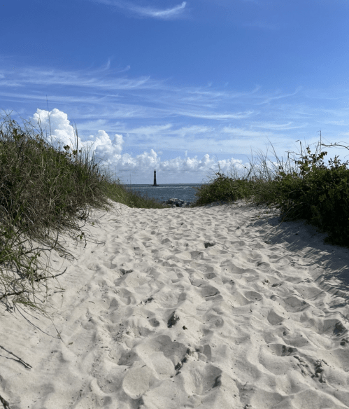 A sandy path leads to the ocean, framed by grass, with a lighthouse visible in the distance under a blue sky.