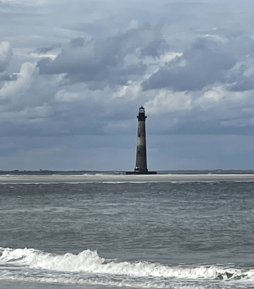A tall lighthouse stands on a sandy shore, surrounded by water under a cloudy sky. Waves gently lap at the beach.