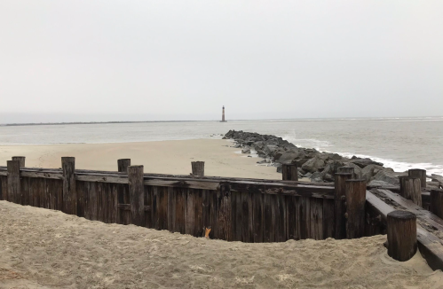 A sandy beach with wooden structures and a distant lighthouse under a cloudy sky.