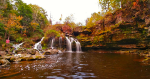 A serene waterfall cascades into a calm pool, surrounded by autumn foliage and rocky cliffs.