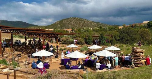 Outdoor event with guests seated under umbrellas, surrounded by greenery and mountains, under a partly cloudy sky.