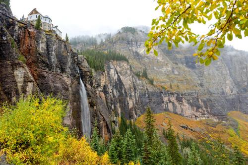 A waterfall cascades down a rocky cliff, surrounded by autumn foliage and a house perched above in a misty landscape.