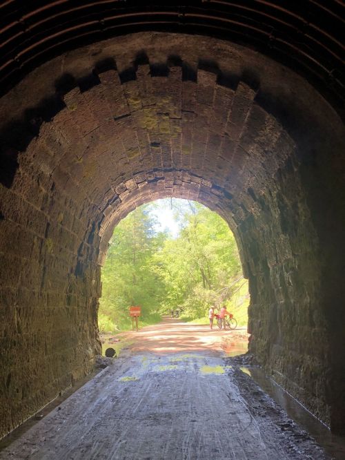 View from inside a stone tunnel, leading to a bright, green landscape with people walking and biking outside.