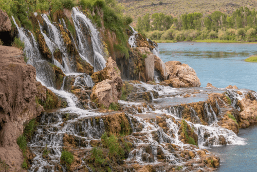 A scenic view of cascading waterfalls over rocky terrain, flowing into a calm river surrounded by greenery.