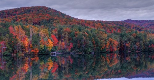 A serene lake reflecting vibrant autumn foliage on surrounding hills under a cloudy sky.
