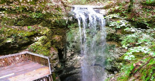 A serene waterfall cascading over rocks, surrounded by lush greenery and a wooden viewing platform.