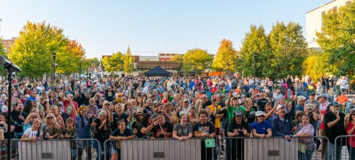 A large crowd of people at an outdoor event, with trees and buildings in the background, enjoying the festivities.