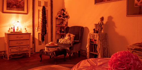 Cozy, warmly lit room with vintage furniture, a plush chair, and decorative dolls on shelves.