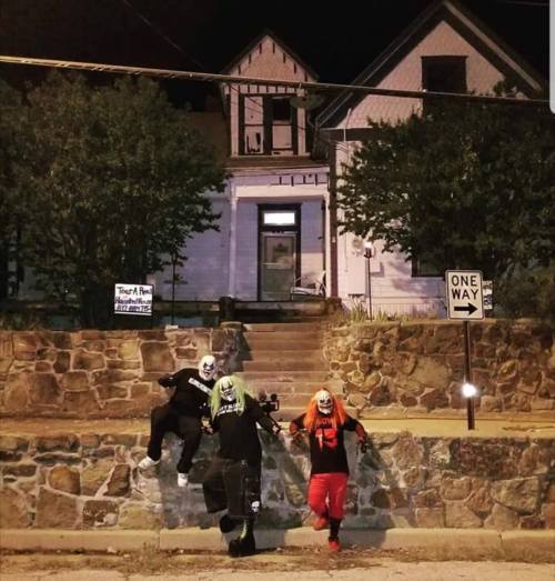 Three people in clown costumes pose in front of a dark, old house at night, with a "One Way" sign nearby.