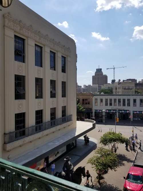 View of a city street with a historic building, people walking, and modern skyscrapers in the background under a blue sky.