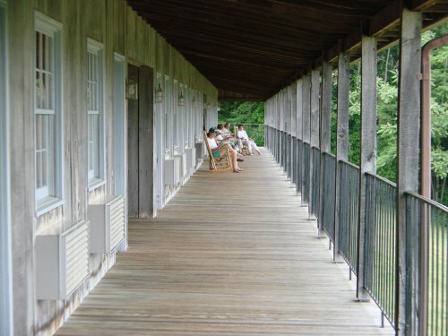 A wooden porch with several people sitting in rocking chairs, surrounded by greenery and natural light.