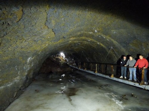 A dark, rocky cave with a wooden bridge and three people standing on it, illuminated by faint lights.