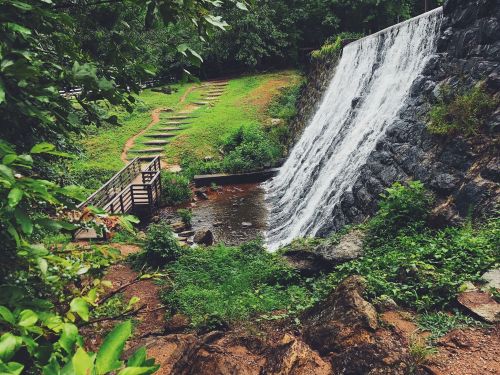 A serene waterfall cascades over rocks, surrounded by lush greenery and a wooden bridge in a tranquil natural setting.