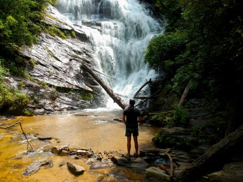 A person stands near a waterfall, surrounded by lush greenery and rocky terrain, with water cascading down the rocks.